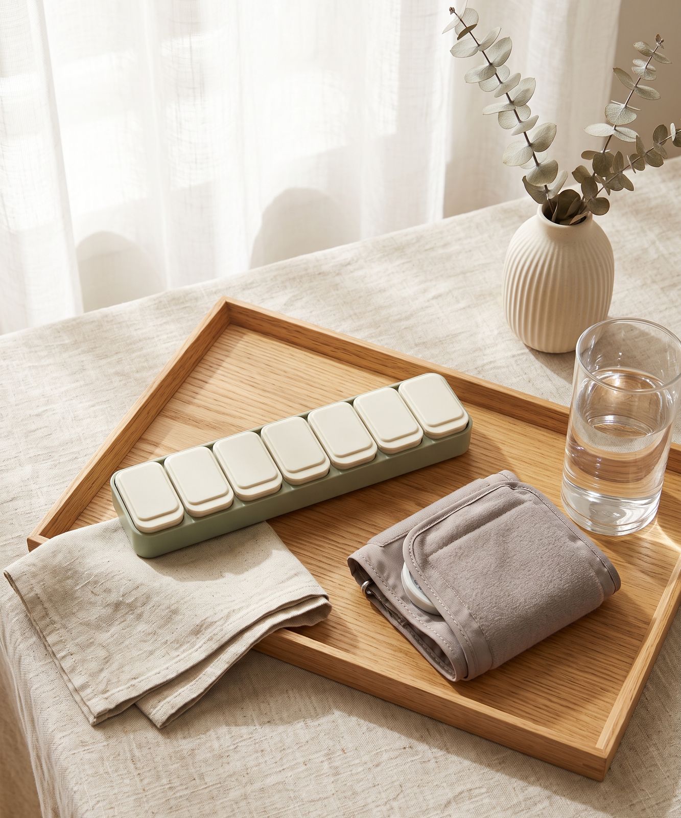 Weekly medication organizer, blood pressure cuff and eucalyptus on a wooden tray in morning light